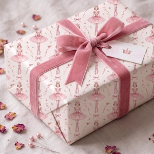 Op de afbeelding: Een rechthoekige geschenkdoos verpakt in wit papier met een herhalend patroon van ballerina's in roze tutu's en kronen. De doos is gestrikt met een breed roze fluwelen lint en bevat een wit rechthoekig cadeaulabel met een kleine kroonafbeelding.