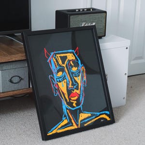 Könnte beinhalten: Gerahmter Kunstdruck mit einer farbenfrohen Illustration einer Person mit Hörnern, roten Lippen und blauen und gelben Gesichtszügen. Das Kunstwerk ist vor einem schwarzen Hintergrund platziert und in einem schwarzen Rahmen gerahmt. Die Kunst lehnt an einer Wand.