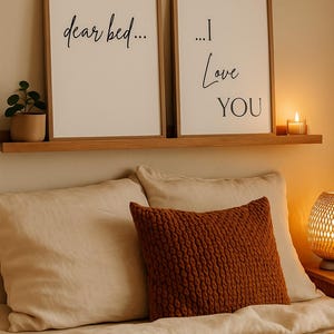 Könnte beinhalten: Zwei gerahmte Kunstdrucke mit den Schriftzügen "dear bed..." und "...I Love YOU" in schwarzer Schrift. Eine kleine Topfpflanze und eine brennende Kerze stehen auf einem Holzregal über einem Bett mit beiger Bettwäsche und einem strukturierten rostfarbenen Kissen.