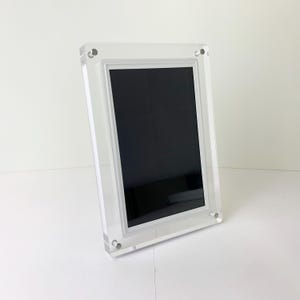 May include: A clear acrylic photo frame with a black insert. The frame is rectangular with rounded corners and is held together with small silver screws. The frame is standing upright on a white surface.