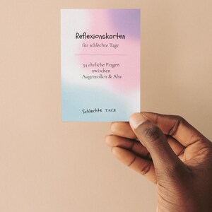 May include: A hand holds a rectangular card with a blue, pink, and purple gradient background. The card displays German text, including "Reflexionskarten" and "für schlechte Tage". The card's design is simple and modern.