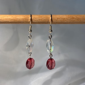 May include: A pair of dangle earrings with silver-colored hooks. Each earring features a clear, faceted bead, a silver-colored connector, and a deep red, oval-shaped bead. The earrings are displayed against a light background.
