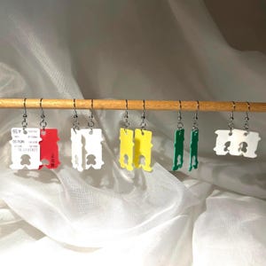Può includere: Orecchini fatti a mano con etichette di sacchetti per il pane riciclate in vari colori, tra cui rosso, bianco, giallo e verde. Gli orecchini sono appesi a ganci d'argento su un'asta di legno.