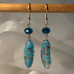May include: A pair of dangle earrings with silver-colored hooks. Each earring features a faceted, dark teal bead above a clear, oval-shaped bead with a turquoise and gold swirl design. The earrings are suspended from a wooden rod.