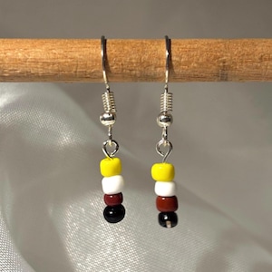 Könnte beinhalten: Ein Paar Perlenohrringe mit silberfarbenen Haken. Jeder Ohrring hat einen Stapel aus fünf Perlen: gelb, weiß, kastanienbraun und schwarz. Die Ohrringe hängen an einem Holzstab vor einem hellen Hintergrund.