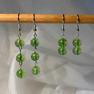May include: Four pairs of green beaded earrings with silver-colored hooks are displayed on a wooden dowel. Two pairs have three beads each, and two pairs have two beads each. The beads are a translucent, light green color.
