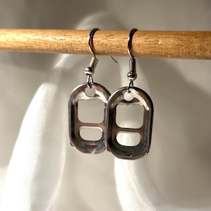 Op de afbeelding: Een paar zilverkleurige oorbellen gemaakt van gerecyclede blikjes. De oorbellen hangen aan zilverkleurige oorhaken. De blikjes hebben een rechthoekige vorm met een centrale opening. De oorbellen worden op een houten deuvel getoond.