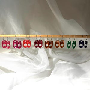 Może przedstawiać: Kolekcja kolorowych kolczyków wykonanych z przetworzonych zawleczek od puszek. Kolczyki są w różnych odcieniach różu, czerwieni, pomarańczy, zieleni i niebieskiego, każdy z unikalnym metalicznym połyskiem. Wiszą na drewnianym drążku na tle białej tkaniny.