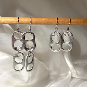 Peut inclure: Boucles d'oreilles argentées fabriquées à partir de languettes de canettes recyclées. Les boucles d'oreilles sont suspendues à des crochets argentés. Deux paires de boucles d'oreilles sont visibles, une avec deux languettes reliées et une autre avec une seule languette.