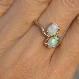May include: A gold ring featuring two oval opals. The opals have a milky white color with flashes of iridescent green. The ring's band has a delicate, flowing design.