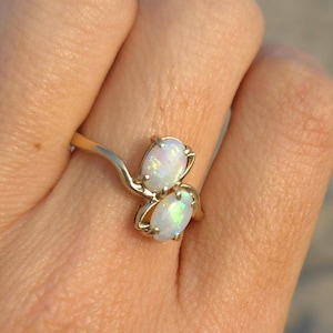 May include: A gold ring featuring two oval-shaped opals. The opals have a pearlescent white color with hints of iridescent hues. The ring is set against a blurred beach background.