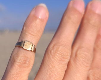 May include: A gold-toned signet ring featuring a flat, rectangular face with a textured surface. The band is smooth and tapers slightly toward the back, showcasing a polished metallic finish that reflects light across its surface.