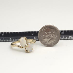 May include: A gold ring featuring two oval-shaped opals. The ring has a swirling design and is next to a U.S. dime for size comparison. The opals have a milky white appearance.