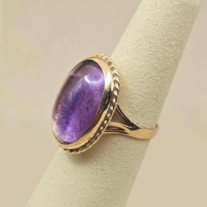 May include: A gold ring featuring a large, oval-shaped amethyst gemstone. The amethyst has a deep purple hue with lighter internal patterns. The ring's setting has a twisted rope design around the gemstone.