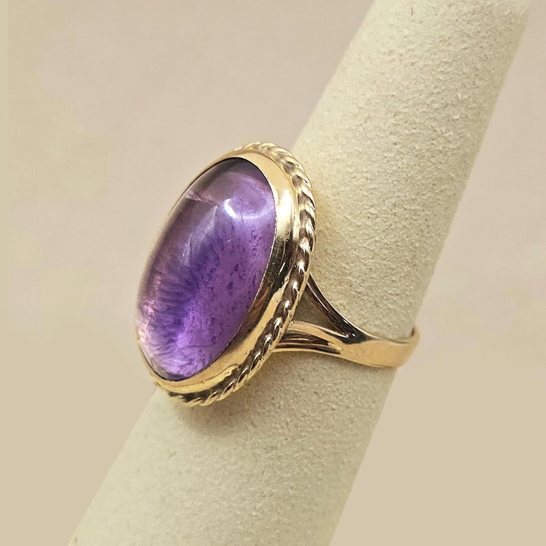 May include: A gold ring featuring a large, oval-shaped amethyst gemstone. The amethyst has a deep purple hue with lighter internal patterns. The ring's setting has a twisted rope design around the gemstone.