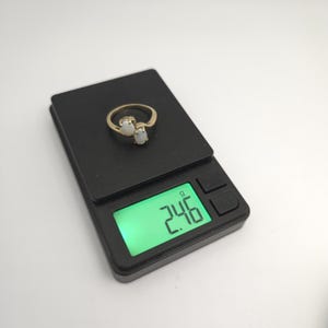 May include: A gold ring with two oval-shaped, white gemstones. The ring is on a black digital scale, which displays a weight of 2.46 grams.