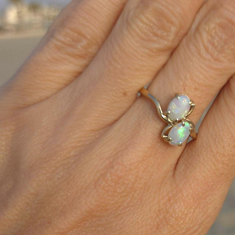 May include: A gold ring featuring two oval opals. The opals have a milky white color with flashes of iridescent green. The ring's band has a delicate, flowing design.