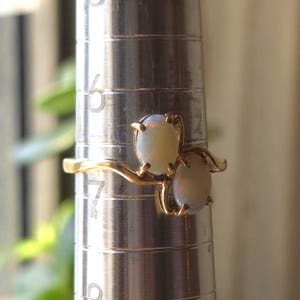 May include: A gold ring featuring two oval opals. The opals have a milky white color with iridescent flashes. The ring is on a silver ring sizer, showing the size.