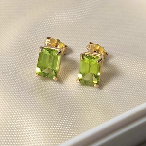 May include: A pair of stud earrings featuring rectangular, emerald-cut lime green gemstones set in gold metal prongs. Each earring has a post back mechanism, showcasing the vibrant green color and faceted surface of the stones in a classic design.