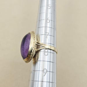 May include: A gold ring with a large, oval, purple gemstone. The ring has a textured, rope-like border around the stone. The ring is being measured on a silver ring sizer.