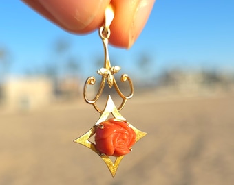 May include: A gold pendant featuring a carved coral rose set within a star-shaped frame. The pendant has an ornate design with curved accents and a small loop for a chain. The background is a blurred view of a sunny outdoor scene.
