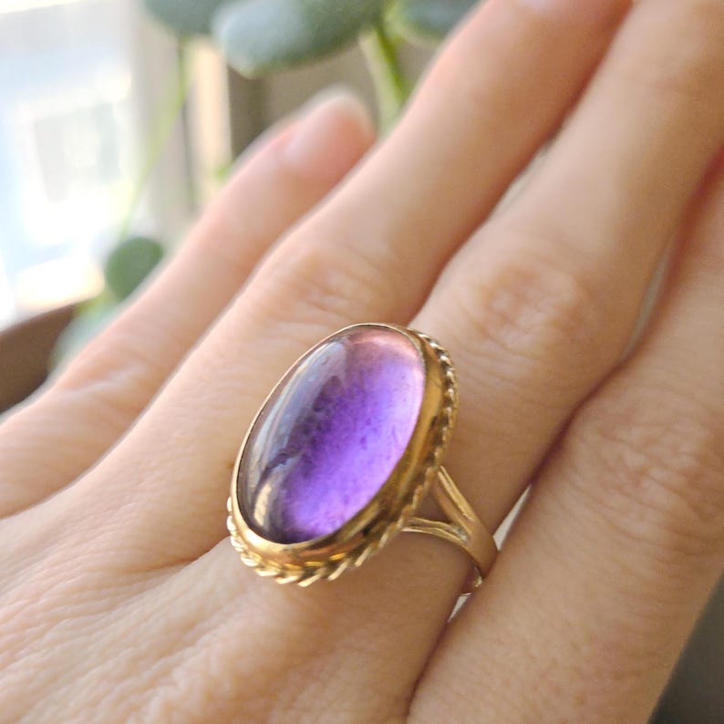May include: A gold ring featuring a large, oval-shaped purple gemstone. The gemstone has a gradient of colors, transitioning from a lighter pinkish hue to a deep purple. The ring's band and setting are gold, with a rope-like texture around the gemstone.