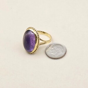 May include: A gold-toned ring with an oval, deep purple gemstone. The ring's band has a split design and a textured rope-like border around the gemstone. A U.S. dime is next to the ring for size comparison.