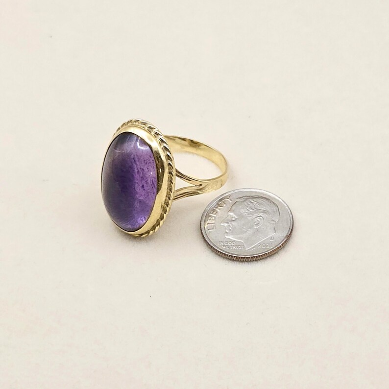 May include: A gold-toned ring with an oval, deep purple gemstone. The ring's band has a split design and a textured rope-like border around the gemstone. A U.S. dime is next to the ring for size comparison.