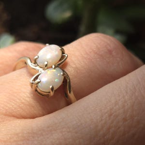 May include: A gold ring featuring two oval-shaped opals. The opals have a milky white base color with iridescent flashes of color. The ring's band and setting are a polished gold color, creating a classic and elegant design.