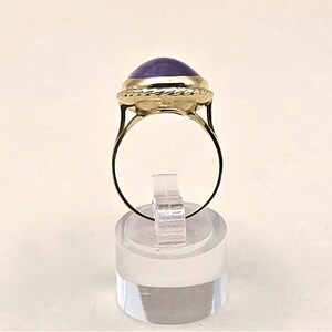 May include: A gold ring featuring a large, oval, purple gemstone. The gemstone is set in a gold bezel with a twisted rope design around it. The ring is displayed on a clear, cylindrical stand.