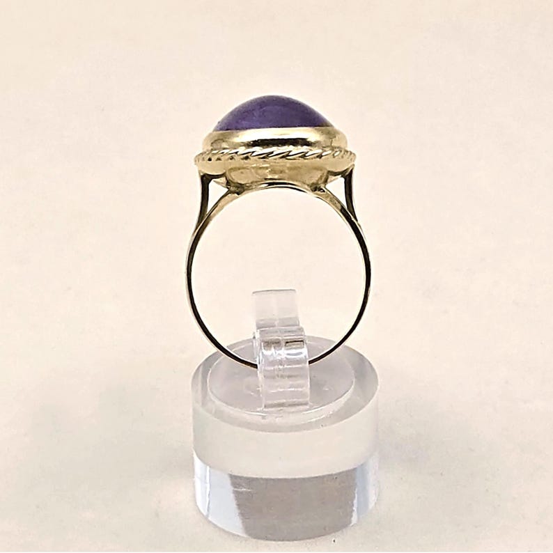 May include: A gold ring featuring a large, oval, purple gemstone. The gemstone is set in a gold bezel with a twisted rope design around it. The ring is displayed on a clear, cylindrical stand.
