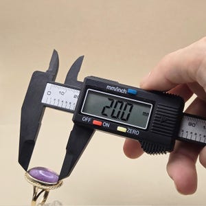 May include: A digital caliper measuring a gold ring with a purple stone. The digital display reads 20.0 mm. The ring has a textured gold band and a round, polished purple stone.