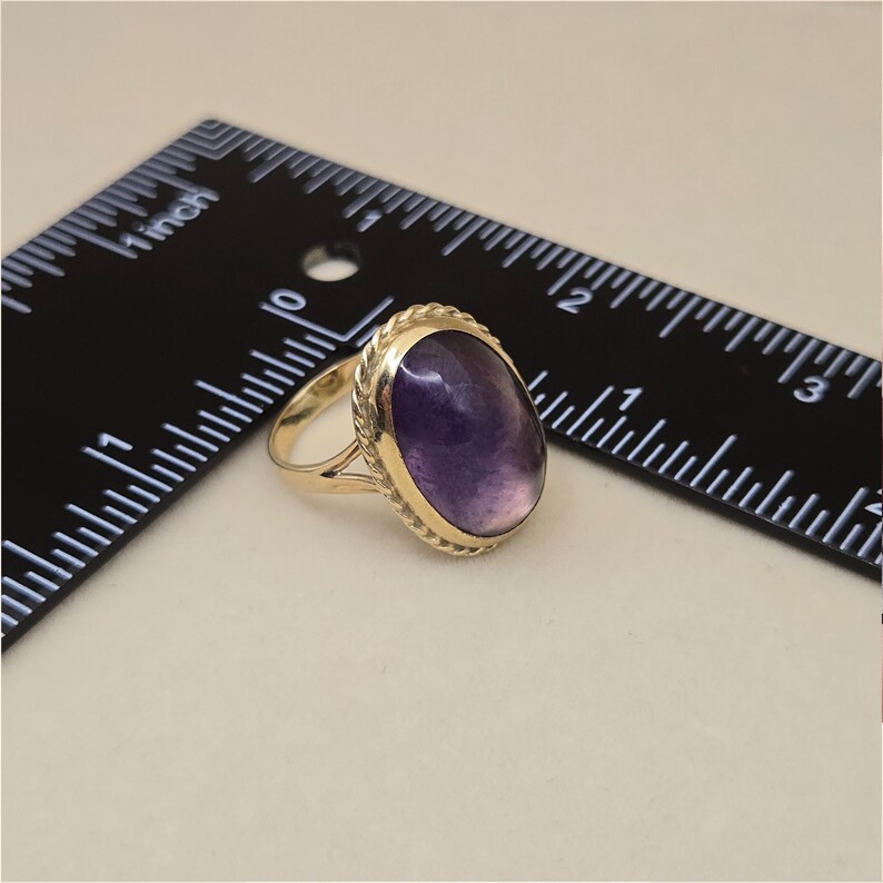 May include: A gold ring with a large, oval, purple gemstone. The gemstone is set in a gold bezel with a rope-like border. The ring is next to a black ruler with inch measurements.