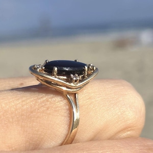 May include: A gold ring featuring a smooth, oval-shaped black stone set in the center. The metal band has a split design near the setting, which is accented with small, clear, sparkling stones embedded along the edges of the gold frame.