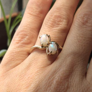 May include: A gold ring featuring two oval opals. The opals have a milky white base with flashes of iridescent color. The ring's band curves gracefully, holding the stones in a secure setting. The ring is shown on a person's finger.