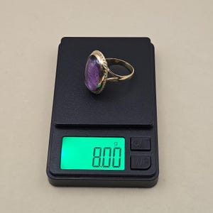 May include: A gold ring with a large, oval-shaped purple gemstone. The ring is on a black digital scale, which displays a weight of 8.00 grams. The gemstone is set in a decorative gold bezel.