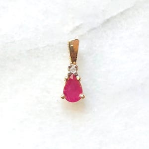 May include: A gold pendant featuring a pear-shaped red gemstone at the base, topped with a small, clear, round-cut stone set in a gold prong setting. The pendant includes a gold bail at the top for attachment to a chain or necklace.