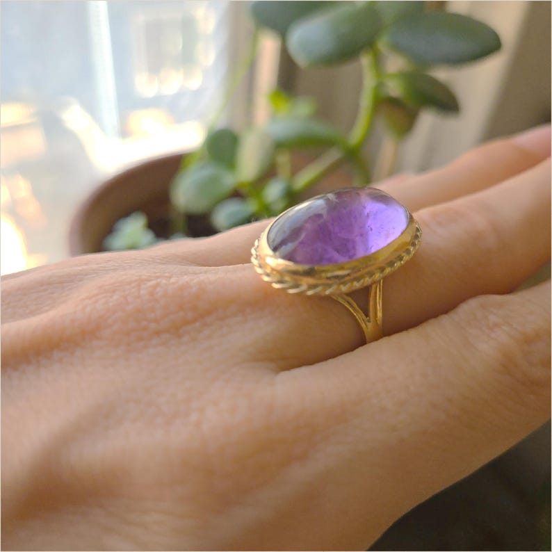 May include: A gold ring featuring a large, oval-shaped purple gemstone. The gemstone is set in a gold band with a braided rope design around the stone. The ring is on a person's hand.