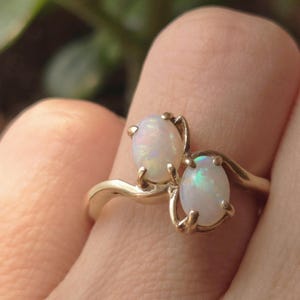 May include: A gold ring featuring two oval-shaped opals. The opals have a milky white base with flashes of iridescent color. The ring's band curves gracefully, and the opals are held in place by delicate prongs.