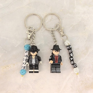 May include: Keychains featuring Lego-style figures wearing hats and jackets. One keychain has the name "LOGAN" spelled out with white letter beads and blue accents. Another keychain has the name "EDWARD" spelled out with white letter beads and crystal accents.