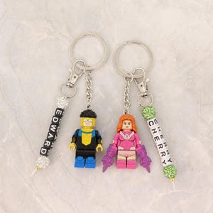 May include: Two keychains with Lego-style figures. One keychain has a yellow and black figure, the other has a pink figure. Both keychains have beaded accents and spell out names: "EDWARD" and "CHERRY".
