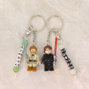 May include: Two keychains featuring Lego figures of characters with lightsabers. One figure has a tan outfit and a blue lightsaber, the other has a black outfit and a red lightsaber. Each keychain has a name spelled out in beads.
