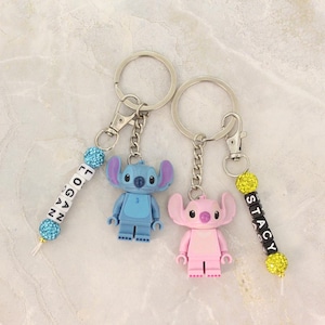 May include: Two keychains featuring cartoon character figures, one blue and one pink, with personalized beaded name tags. The keychains have silver metal rings and clasps. The name tags spell out "LOGAN" and "STACY".