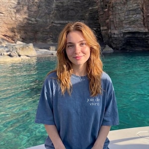 May include: A woman wearing a blue t-shirt with the words "joie de vivre" printed on the front. She is on a boat with a turquoise body of water and a rocky cliff in the background.