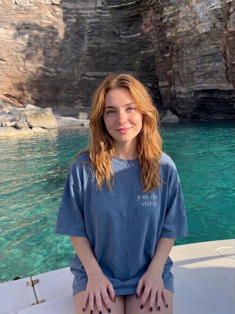 May include: A woman wearing a blue t-shirt with the words "joie de vivre" printed on the front. She is on a boat with a turquoise body of water and a rocky cliff in the background.