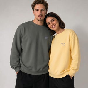 May include: Two people wearing matching sweatshirts. One sweatshirt is a muted green, the other is yellow. Both sweatshirts have the text "joie de vivre" in a small, purple font on the chest. They are both wearing black pants.