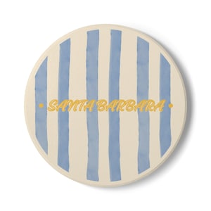Pode incluir: Base para copos redonda com fundo creme e listras verticais azul claro. O texto "SANTA BARBARA" é impresso em amarelo no centro da base.