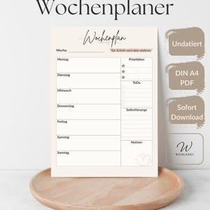 May include: A minimalist weekly planner with a cream-colored background, featuring sections for each day of the week, priorities, to-do lists, and notes. The planner includes the text "Minimalistischer Wochenplaner" and "Wochenplan". It is displayed on a wooden tray.
