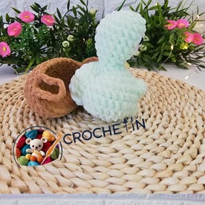 Puede incluir: Un pato de ganchillo y una pequeña cesta en tonos marrón y azul claro, sobre una esterilla tejida. La imagen incluye el texto "CROCHE TIN" y un logotipo con un osito de peluche e hilo.