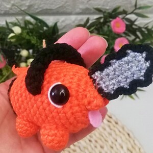 Puede incluir: Una criatura de ganchillo naranja hecha a mano con una cuchilla de motosierra negra y pelo negro. La criatura tiene una lengua rosa que sobresale y un gran ojo negro. El animal de ganchillo está sostenido en la mano de una persona.